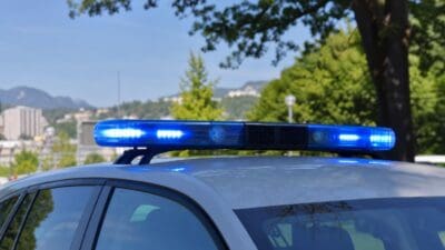 Polizeifahrzeug mit leuchtendem Blaulichtbalken auf dem Dach vor Bäumen und Gebäuden.