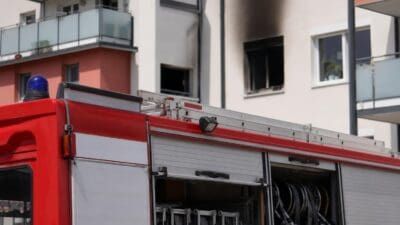 Rotes Feuerwehrfahrzeug mit geöffneten Abteilen vor einem mehrgeschossigen Wohnhaus mit Brandspuren
