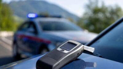 Ein Alkoholtestgerät liegt auf der Motorhaube eines blauen Polizeifahrzeugs vor einem Streifenwagen mit Blaulicht.