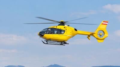 Gelber Hubschrauber fliegt vor blauem Himmel über einer Hügellandschaft.