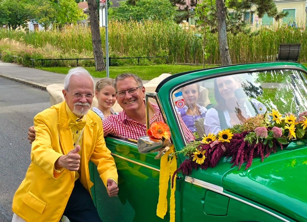 Ein Mann im gelben Anzug posiert neben einem blumengeschmückten grünen Auto mit Menschen darin.