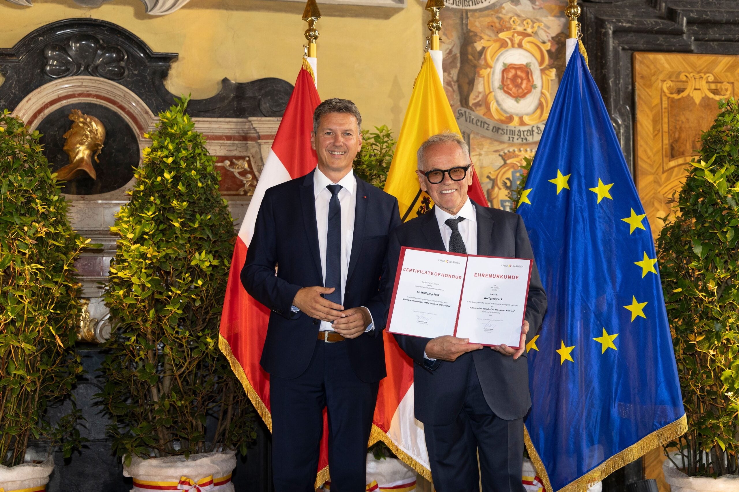 Zwei Männer in dunklen Anzügen präsentieren Ehrenurkunden vor österreichischer und EU-Flagge.