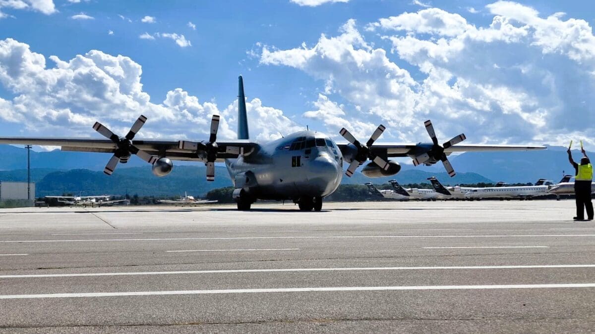 Großes graues Transportflugzeug mit vier Propellern auf dem Flughafenvorfeld, ein Mitarbeiter in Warnweste dirigiert es mit Leuchtstäben.