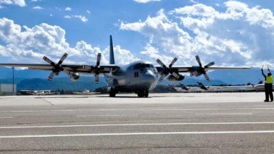 Großes graues Transportflugzeug mit vier Propellern auf dem Flughafenvorfeld, ein Mitarbeiter in Warnweste dirigiert es mit Leuchtstäben.