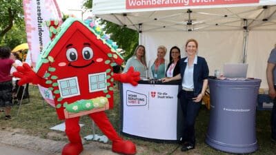 Rotes Hausmaskottchen mit Gesicht und grünem Dach steht vor dem Zelt der Wohnberatung Wien.