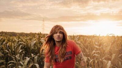 Ein Mann mit langen roten Haaren sitzt auf einem Hocker in einem Maisfeld bei Sonnenuntergang.