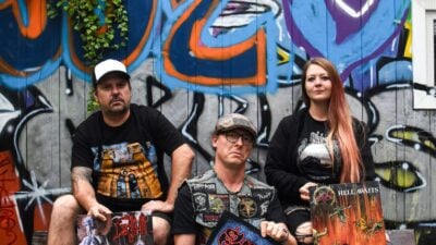 Drei Personen sitzen auf einer Holzbank vor einer bunten Graffiti-Wand und halten Albumcover in den Händen.