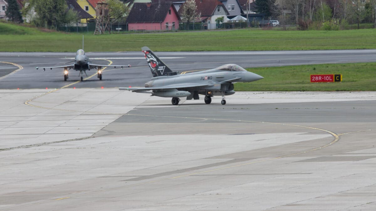 Zwei graue Eurofighter auf der Startbahn eines Flughafens mit Häusern im Hintergrund.