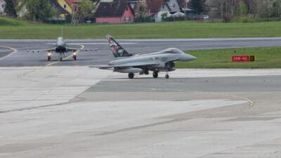 Zwei graue Eurofighter auf der Startbahn eines Flughafens mit Häusern im Hintergrund.