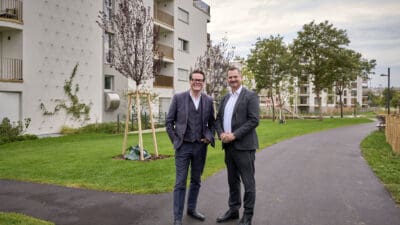 Zwei Männer in Anzügen stehen nebeneinander auf einem Weg in einem Park vor modernen Wohngebäuden.