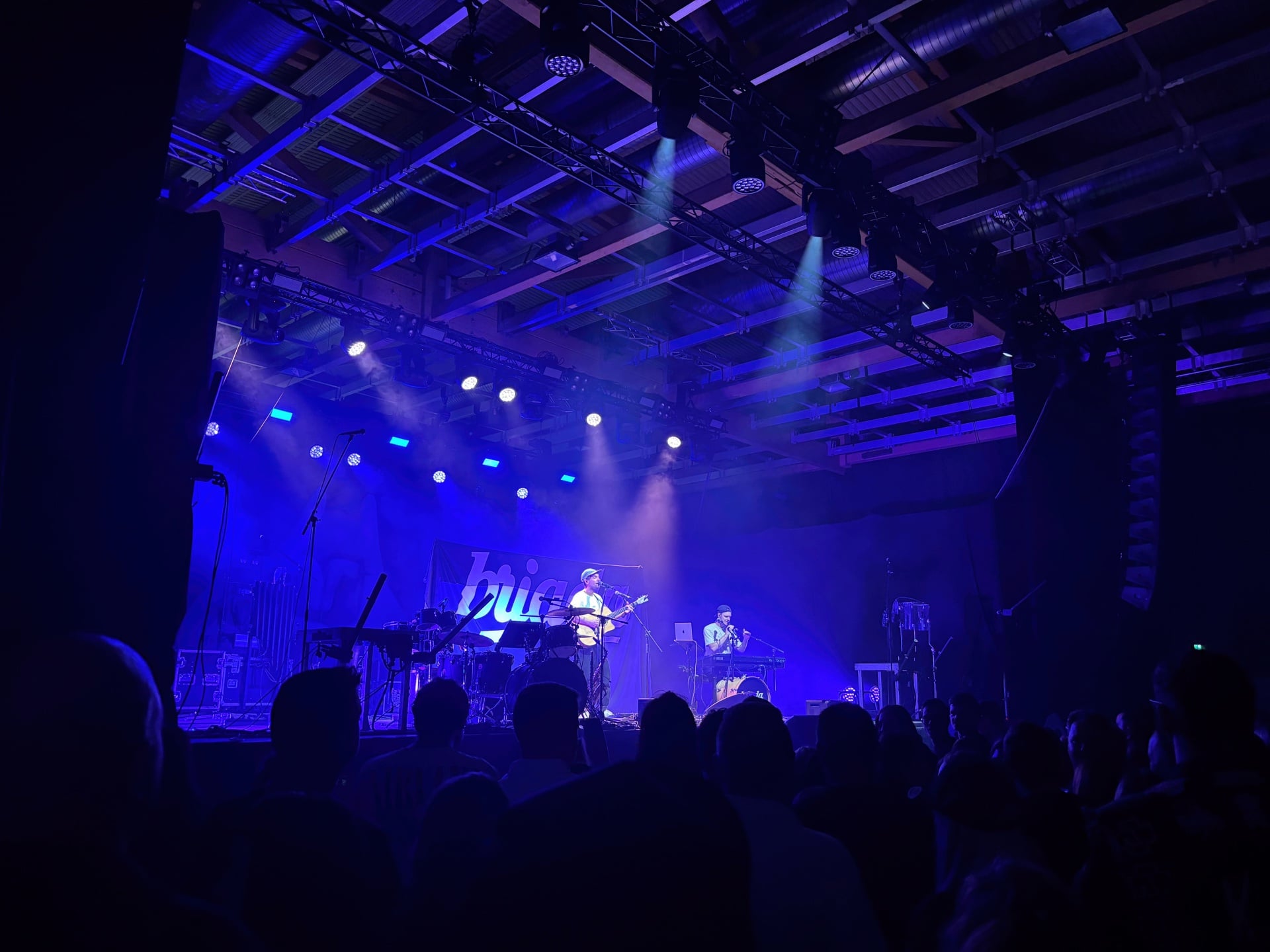 Musiker spielen auf blau beleuchteter Bühne vor dem Publikum in der Messehalle Klagenfurt