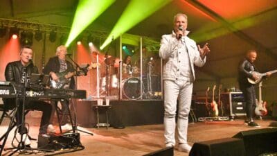 Sänger in weißer Jacke singt auf Bühne mit Band am Keyboard, Bass und Gitarre.