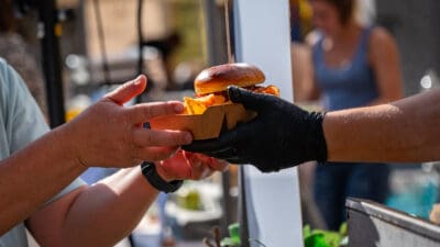 Eine Hand mit schwarzem Handschuh übergibt einen Burger und Pommes in einer braunen Pappbox an eine andere Hand.