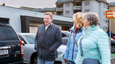 Ein Mann und zwei Frauen gehen an parkenden Autos und Gebäuden vorbei.