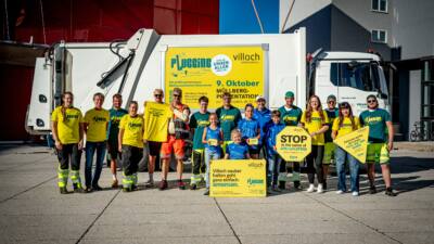 Gruppe von Erwachsenen und Kindern in gelben und grünen Plogging-T-Shirts vor einem weißen Müllwagen mit Bannern.