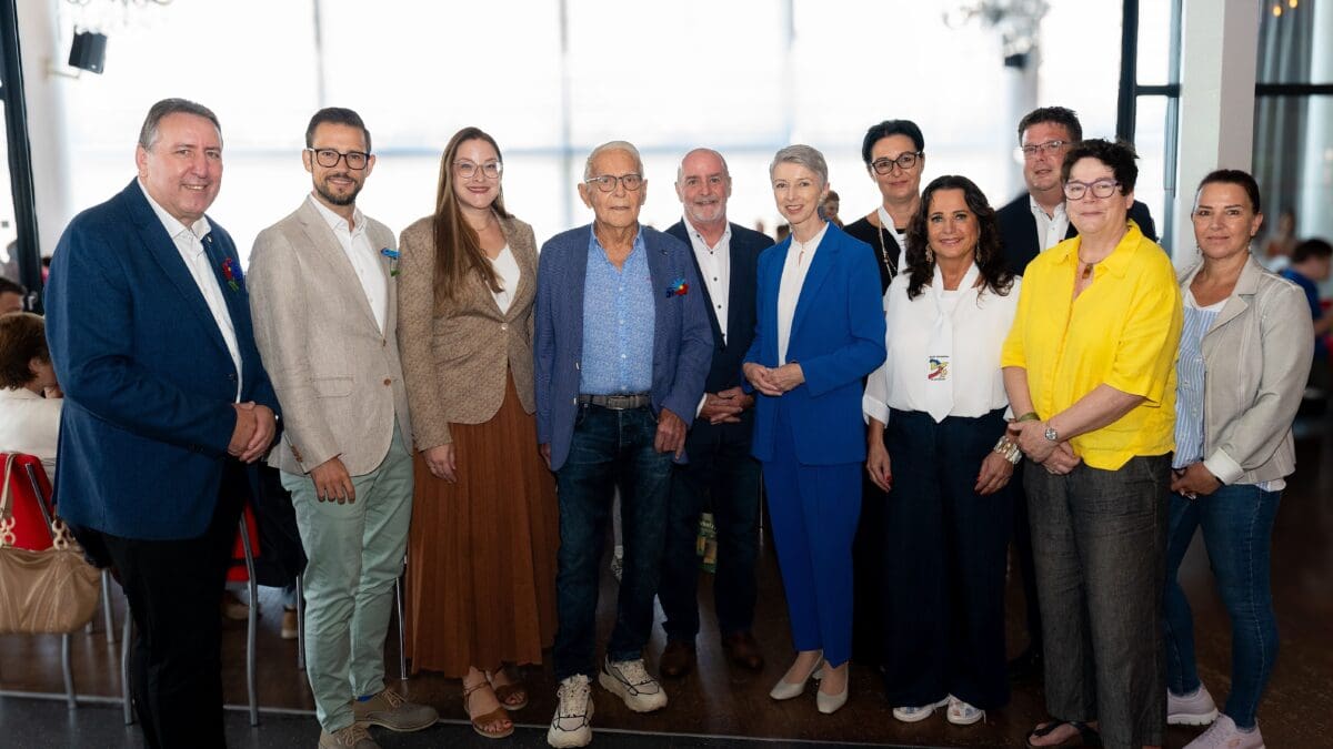 Zehn Erwachsene stehen in einer Reihe für ein Gruppenfoto in einem hellen Saal mit großen Fenstern.