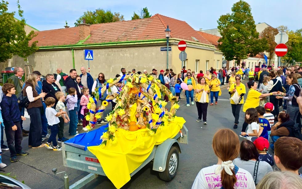 Ein gelb-blau geschmückter Erntedankwagen steht zwischen Zuschauern auf einer Straße.