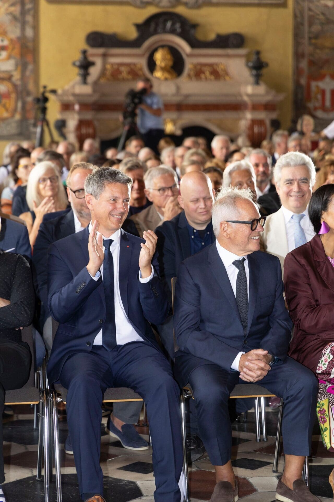 Zwei Männer in dunklen Anzügen sitzen im Publikum und klatschen in einem prunkvollen historischen Saal mit Kamin und Wappen.