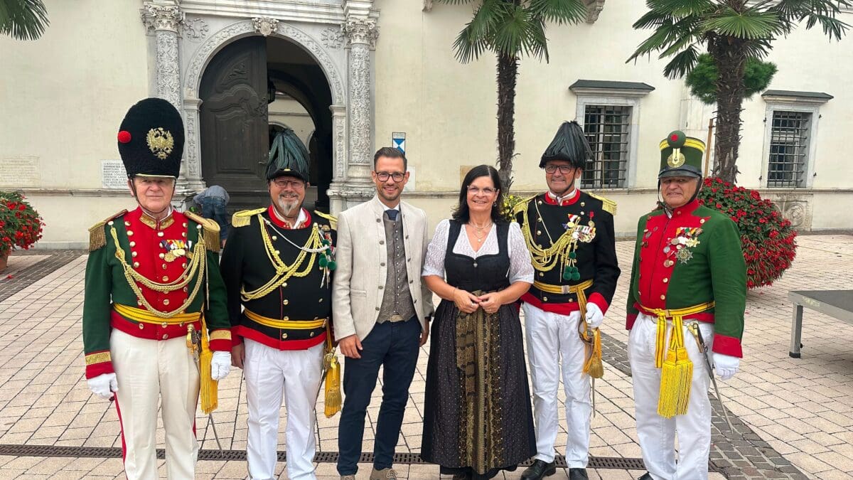 Fünf Gardisten in historischen Uniformen und eine Frau in Tracht vor einem barocken Gebäude mit Palmen