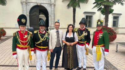 Fünf Gardisten in historischen Uniformen und eine Frau in Tracht vor einem barocken Gebäude mit Palmen