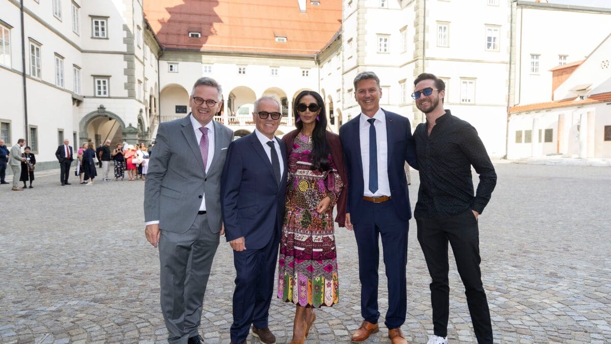 Fünf Personen stehen nebeneinander in einem sonnigen Innenhof mit Kopfsteinpflaster und historischen Gebäuden im Hintergrund.