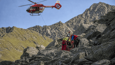 Roter Notarzt-Hubschrauber schwebt über Bergrettern auf felsigem Gelände in den Alpen