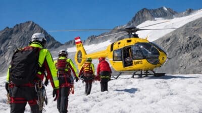 Gruppe von Bergrettern trägt eine Trage über Schnee zu einem gelben ÖAMTC-Helikopter.
