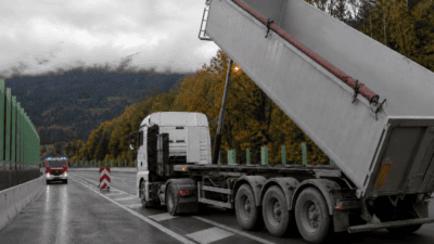 Weißer Sattelschlepper mit hochgestelltem grauem Muldenkipper auf nasser Autobahn, Feuerwehrfahrzeug mit Blaulicht im Hintergrund