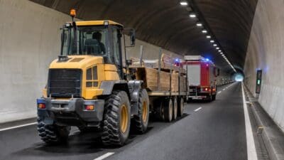 Gelber Radlader mit beladenem Anhänger fährt in einem Tunnel, dahinter rotes Feuerwehrfahrzeug mit Blaulicht.