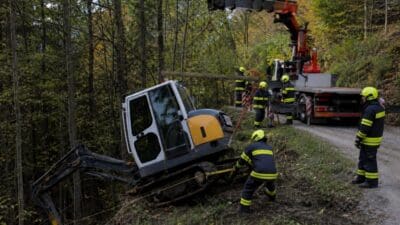 Feuerwehrleute bergen einen umgekippten Bagger mithilfe eines Kranwagens im Wald