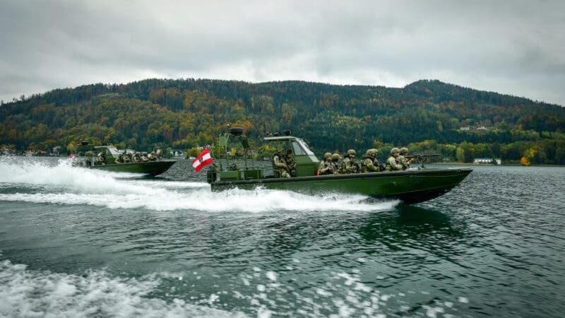 Zwei grüne Militärboote mit Soldaten und österreichischer Flagge fahren auf einem See.