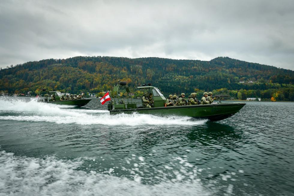 Zwei grüne Militärboote mit Soldaten und österreichischer Flagge fahren auf einem See.