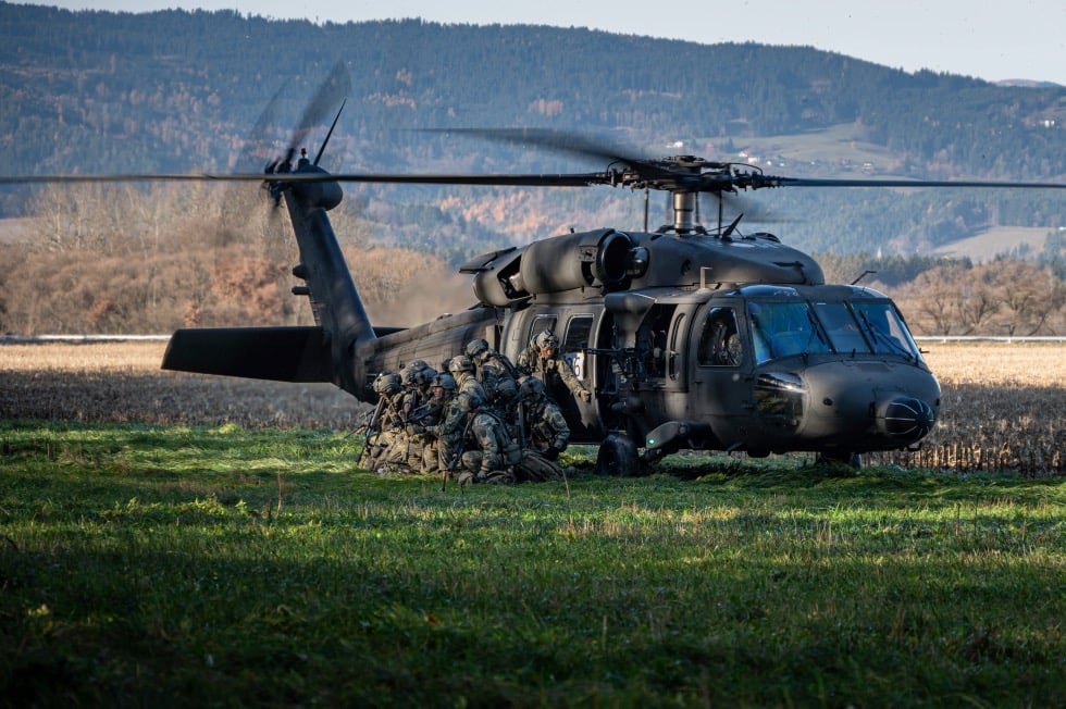 Soldaten in Tarnuniform stehen und sitzen neben einem schwarzen Militärhubschrauber auf einer Wiese vor bewaldeten Hügeln.