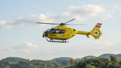 Gelber ÖAMTC-Rettungshubschrauber fliegt über bewaldeten Hügeln unter blauem Himmel.