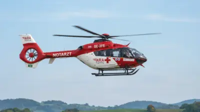 Rot-weißer Notarzthubschrauber fliegt vor blauem Himmel über einer grünen Hügellandschaft.
