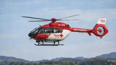 Rot-weißer Notarzthubschrauber der ARA Flugrettung vor bewölktem Himmel über einer bewaldeten Landschaft