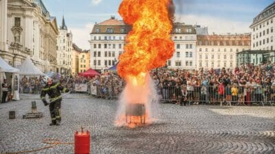 Feuerwehrmann in Schutzkleidung bedient ein langes Werkzeug an einer großen Feuerdemonstration vor einer Menschenmenge