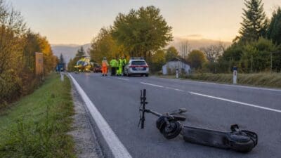 Umgestürzter E-Scooter am Straßenrand mit Einsatzkräften, Polizeiauto und Hubschrauber im Hintergrund