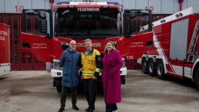 Drei Personen stehen vor einem roten Feuerwehrfahrzeug in einer Fahrzeughalle.
