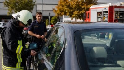 Feuerwehrmann in Schutzkleidung bearbeitet die Tür eines dunklen Autos während ein Polizist daneben steht