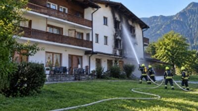 Feuerwehrleute in voller Einsatzkleidung bekämpfen einen Brand an einem Alpengebäude mit Wasserschläuchen.