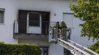 Zwei Feuerwehrleute in Schutzkleidung auf der Plattform einer Drehleiter vor einem verkohlten Balkon
