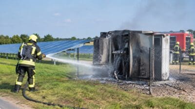 Ein Feuerwehrmann in Schutzkleidung löscht mit einem Wasserschlauch einen Brand an einem verkohlten Elektroschrank neben Solaranlagen.
