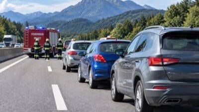Rotes Feuerwehrfahrzeug mit Blaulicht und Feuerwehrleute auf einer Autobahn im Gebirge neben mehreren Autos.