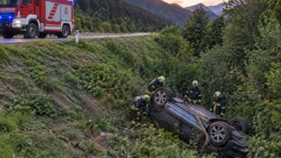 Feuerwehrleute in Einsatzkleidung sichern ein auf dem Dach liegendes Fahrzeug am bewaldeten Hang bei Abendrot.
