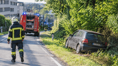 Feuerwehrmann in Schutzkleidung läuft auf Einsatzfahrzeug mit Blaulicht zu, verunglücktes Auto steht am Straßenrand