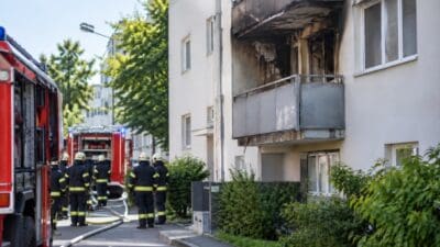 Feuerwehrleute mit Einsatzfahrzeugen vor einem brandbeschädigten mehrstöckigen Wohnhaus