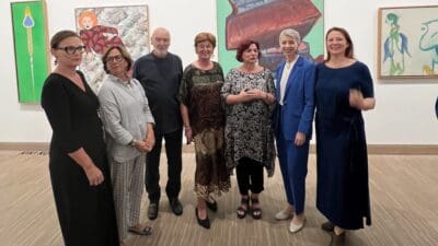 Sechs Frauen und ein Mann posieren in einer Kunstausstellung vor mehreren Gemälden.