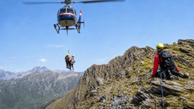 Polizeihubschrauber hebt zwei Personen mit einem Seil aus den Bergen, ein Retter sitzt auf dem Grat.