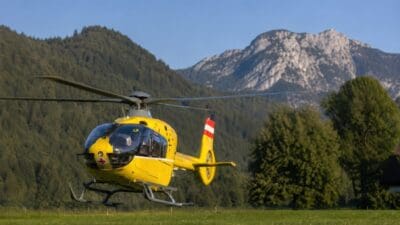 Gelber Rettungshubschrauber mit rotem Kreuz fliegt über grüner Wiese vor Bergpanorama